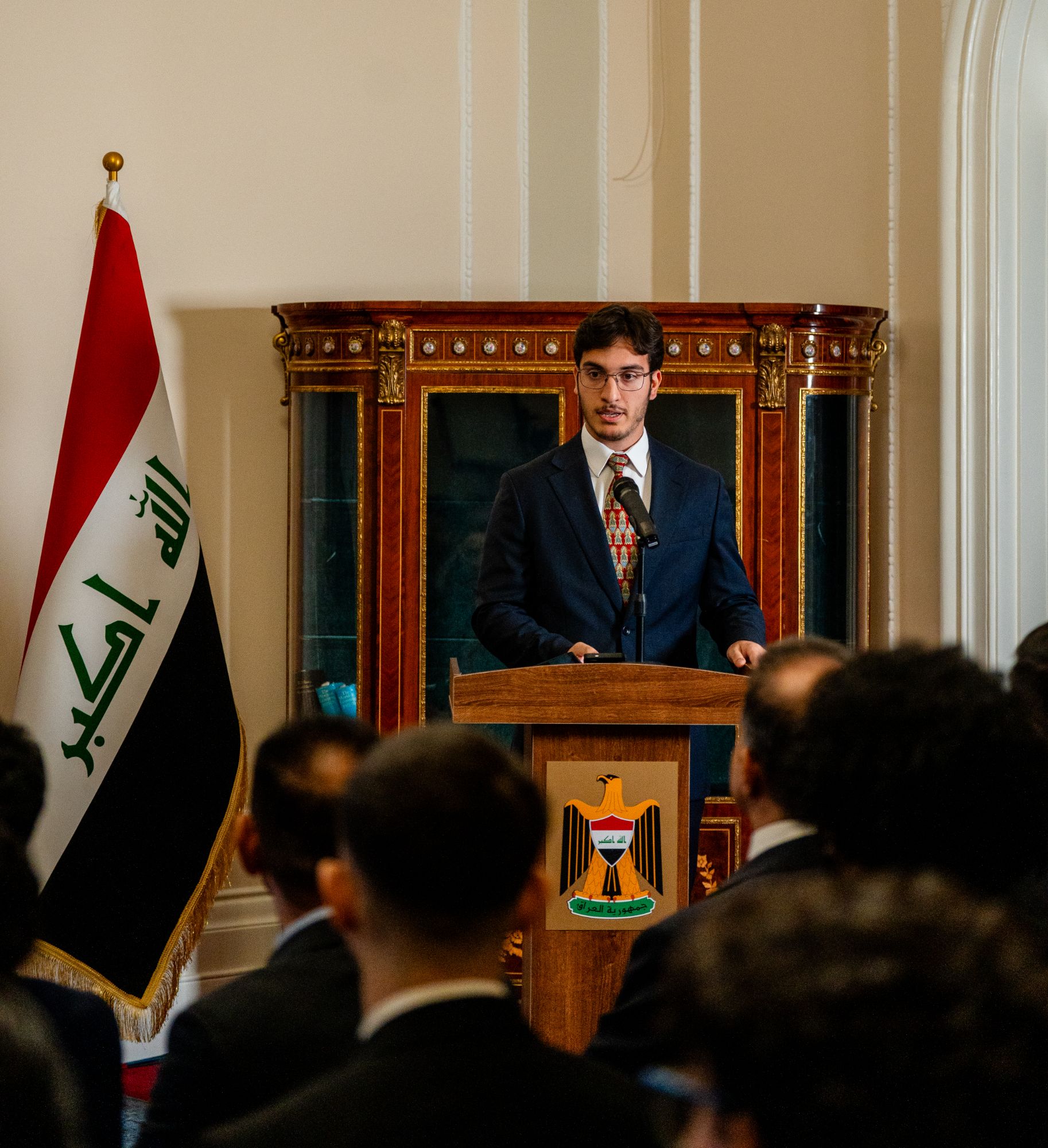 A student speaking at the podium in front of the Iraqi flag